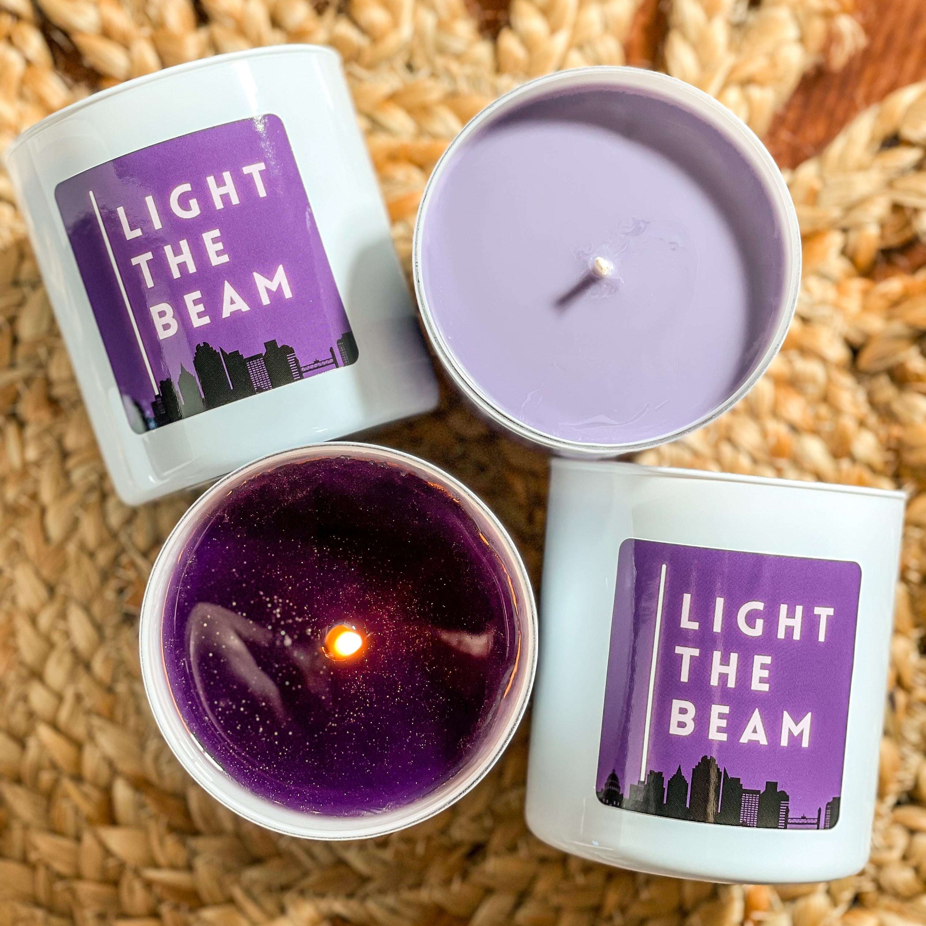 Top view of three Light The Beam candles, one with the lid off showing the purple wax and the flame, and the other two with lids on displaying the label with the brand and logo.